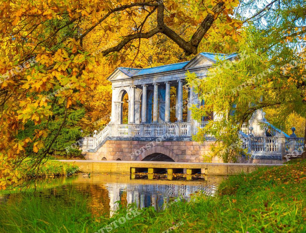 На фотографии изображен Мраморный мост в Екатерининском парке Царского Села. Мост выполнен в классическом стиле и является одной из главных достопримечательностей парка. Осенние деревья окружают мост, окрасив его окружение в насыщенные золотистые и оранжевые тона. Спокойная вода канала отражает архитектуру моста, создавая живописную композицию. Это место притягивает внимание туристов своей исторической красотой и умиротворяющей атмосферой.